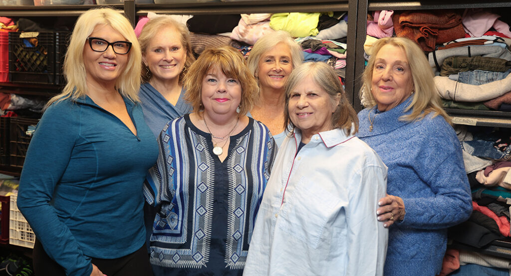 Smiling group portrait of staff members at Wednesday's Child nonprofit. Group is standing in front of a wall with hundreds of clothing donations.