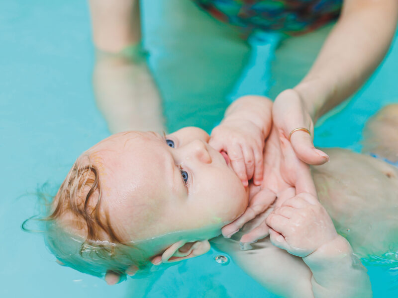 decorative image of baby in a pool with an instructor gently holding the baby's head