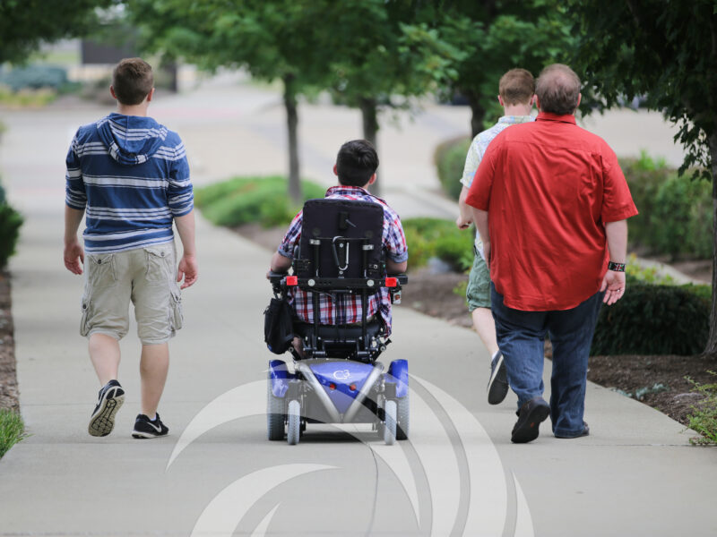 Group of four people, one in wheelchair, walking down a path with their back toward the camera. A white Summit DD swirl logo rises from the bottom of the image.