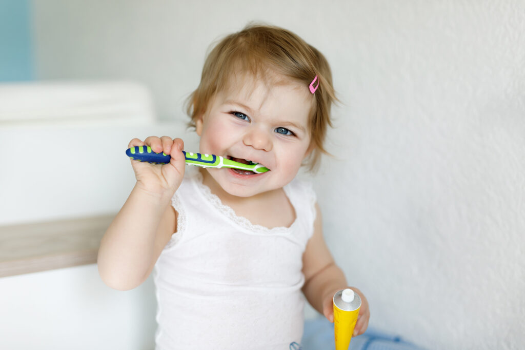Toddler holding toothbrush and toothpast trying to brush her own teeth.