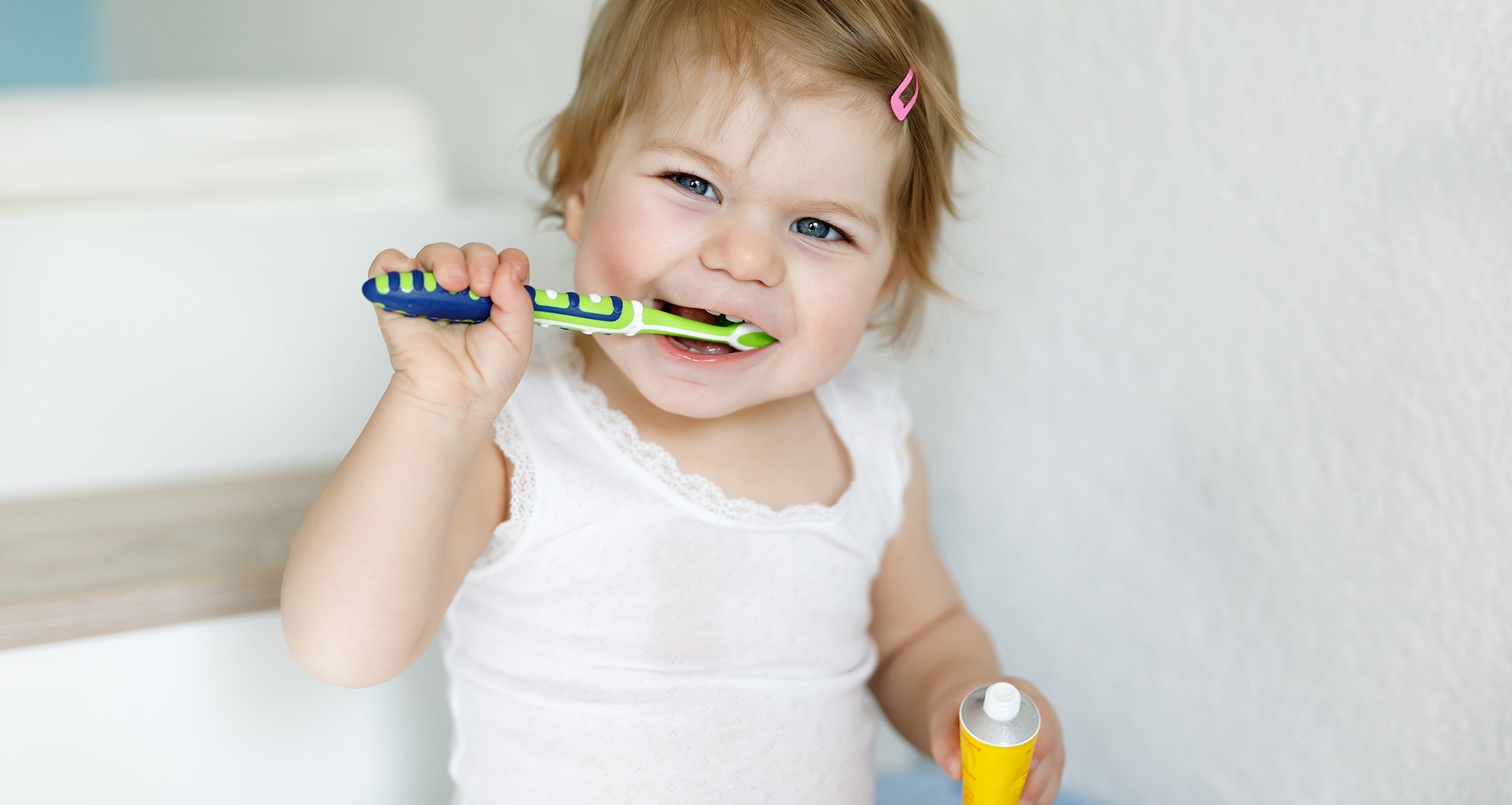 Toddler holding toothbrush and toothpast trying to brush her own teeth.