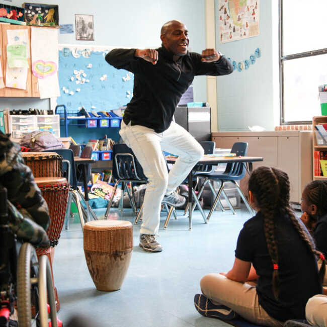 Teaching artist Ssuuna dances as children seated around him watch intently