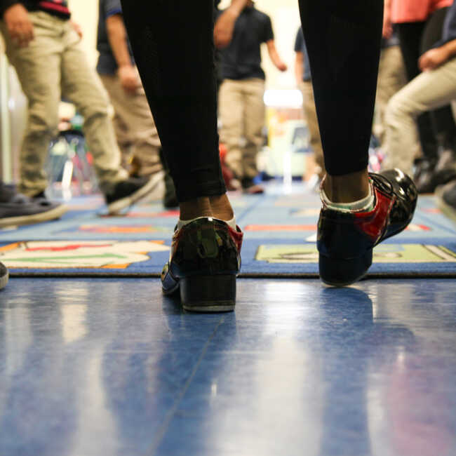 The heel of Quynn's red and black tap shoes strikes the floor in the foreground. In the background, kindergardeners' tennis shoes imitate the dance step.