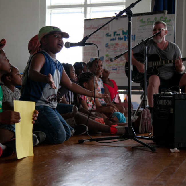 Children kneeling on stage around a microphone and taking turns singing into it with musician Scott Paynter accompanying them on guitar.