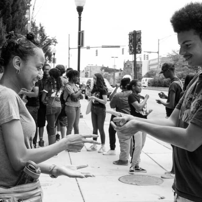 Two high school interns playing Rock, Paper, Scissors on the sidewalk outside of Motor House on North Ave. in Baltimore. A larger group of high school students are gathered in the background.