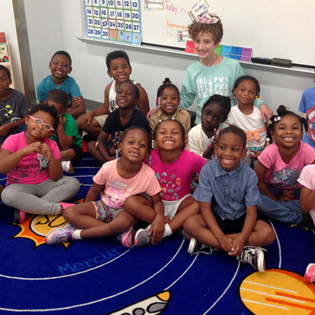The Pre-K class sitting together on a rug depicting the solar system, posing with Alice, a student volunteer.