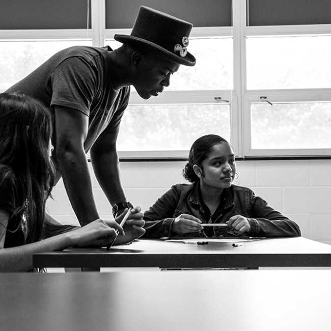 Spoken word artist Femi the Drifish stands at a table where students are seated looking over their work.