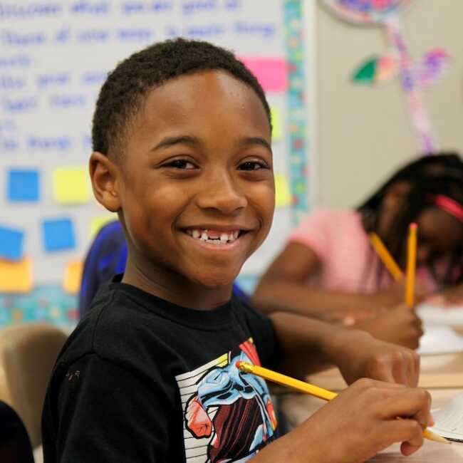 Student sitting at a desk, pencil in hand, looking into the camera and smiling.