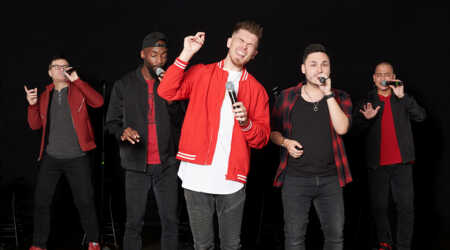 The five members of Ball in the House, dressed in black and red, sing into handheld microphones.