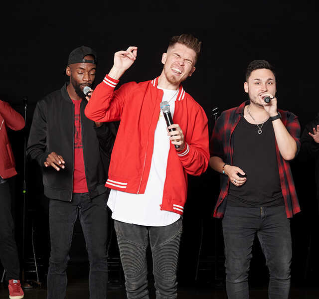 The five members of Ball in the House, dressed in black and red, sing into handheld microphones.