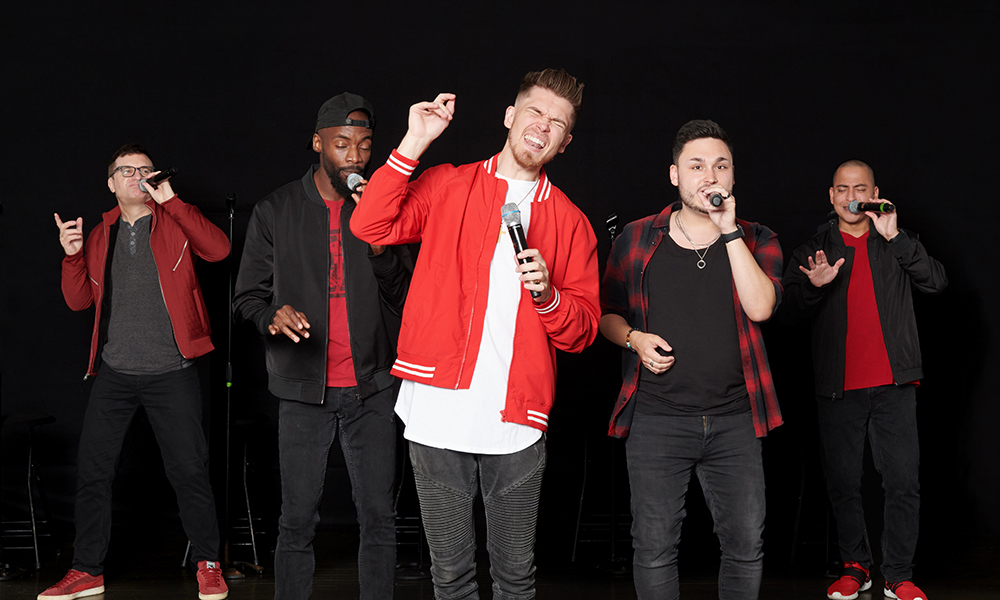 The five members of Ball in the House, dressed in black and red, sing into handheld microphones.