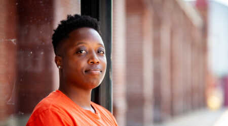 A man in an orange t-shirt with his head turned towards the camera stands in focus in the foreground. The side of a brick building extends into the distance behind him.