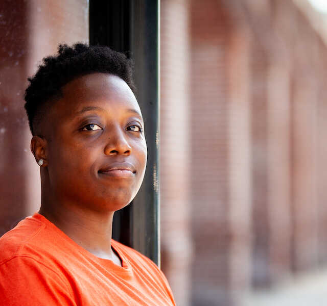A man in an orange t-shirt with his head turned towards the camera stands in focus in the foreground. The side of a brick building extends into the distance behind him.
