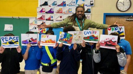 Artist Brandon Richardson poses with students against a wall covered with student-designed sneaker paintings. The students stand in front of the artist and each holds an original sneaker painting with words like "joy" and "unity."