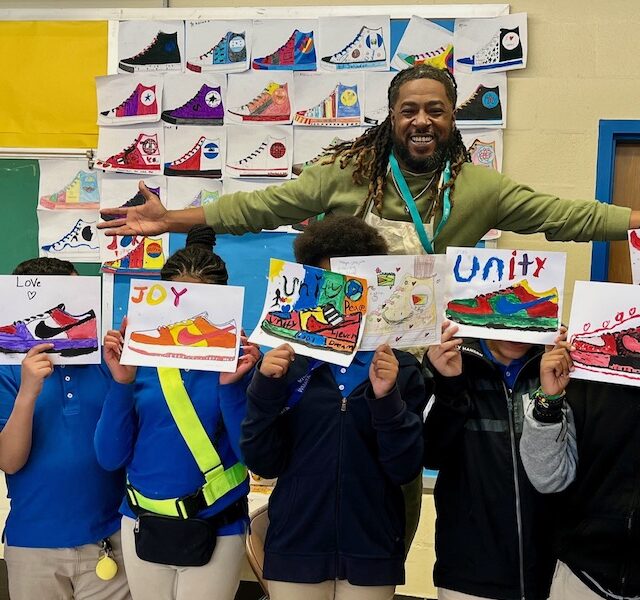 Artist Brandon Richardson poses with students against a wall covered with student-designed sneaker paintings. The students stand in front of the artist and each holds an original sneaker painting with words like "joy" and "unity."