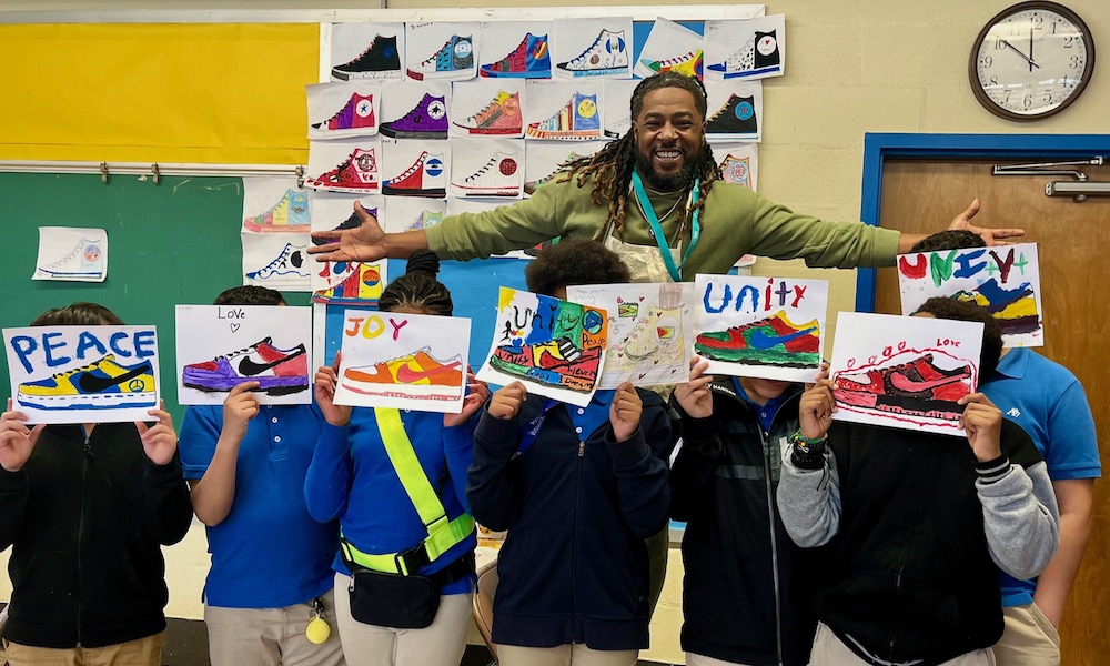 Artist Brandon Richardson poses with students against a wall covered with student-designed sneaker paintings. The students stand in front of the artist and each holds an original sneaker painting with words like "joy" and "unity."