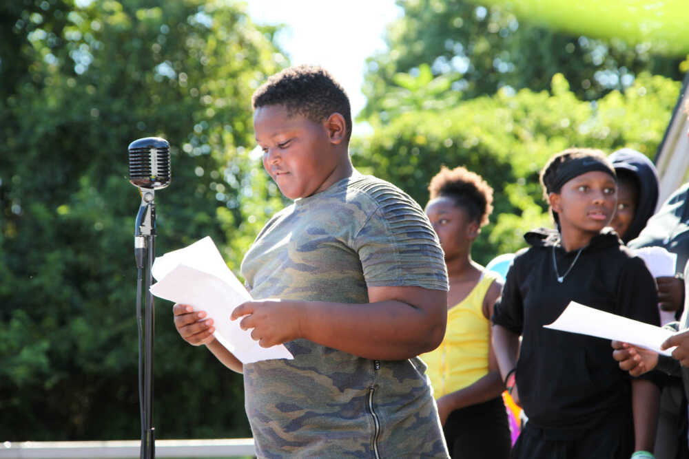 A middle school-aged student ready his poem at the mic onstage with classmates behind him.