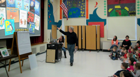 Man stands in front of a projection of book covers as children look on.