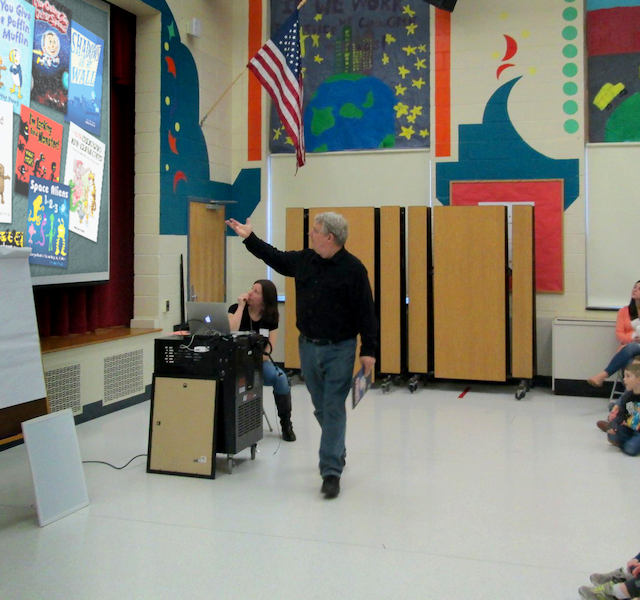Man stands in front of a projection of book covers as children look on.