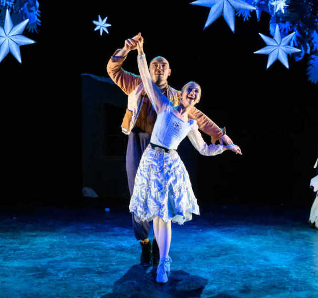 Two dancers on stage with glowing snowflakes and a snow-covered set.
