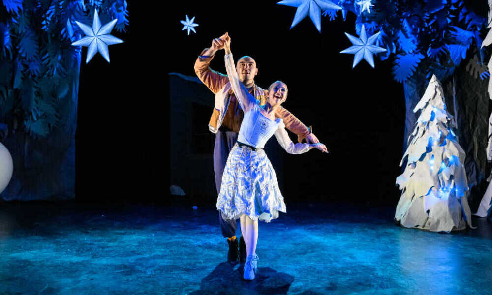 Two dancers on stage with glowing snowflakes and a snow-covered set.