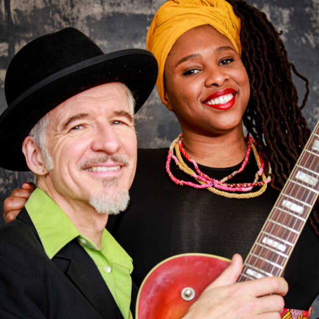 Man wearing a hat, holding a guitar seated with a woman wearing a golden headband and beaded necklace with her arm around his shoulder.