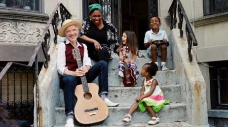 Man with a guitar seated on front steps of a building with a woman and three children.