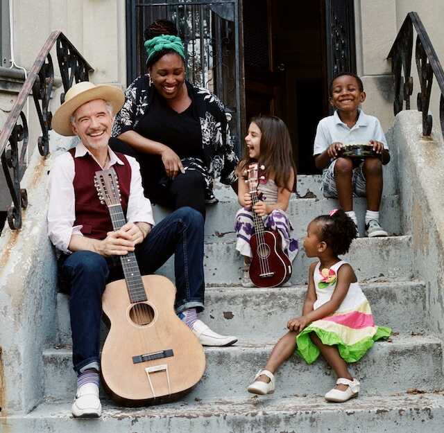 Man with a guitar seated on front steps of a building with a woman and three children.