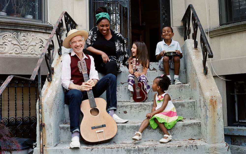 Man with a guitar seated on front steps of a building with a woman and three children.