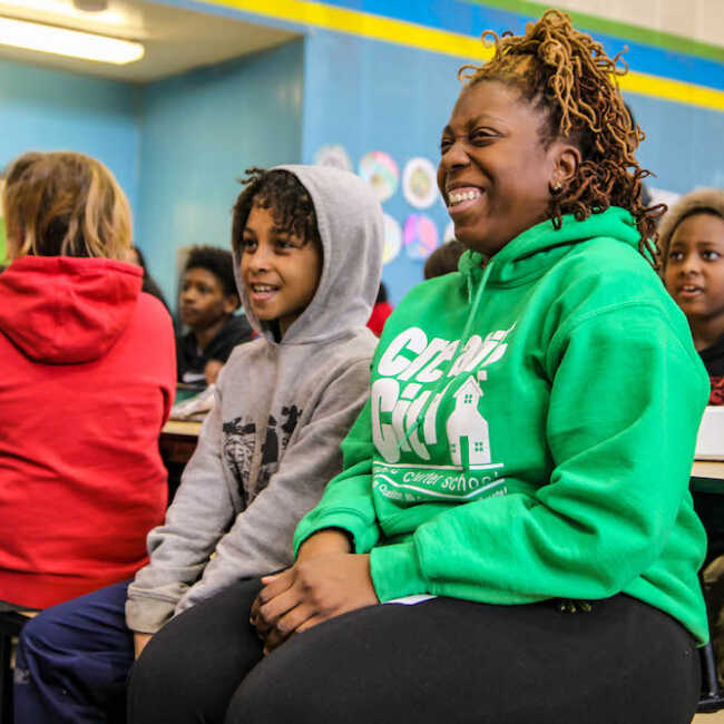 A teacher sits next to a student on bench in a crowded cafeteria. Both are looking ahead and smiling.