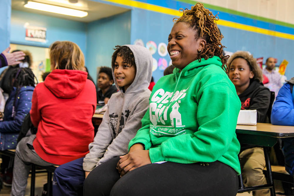 A teacher sits next to a student on bench in a crowded cafeteria. Both are looking ahead and smiling.