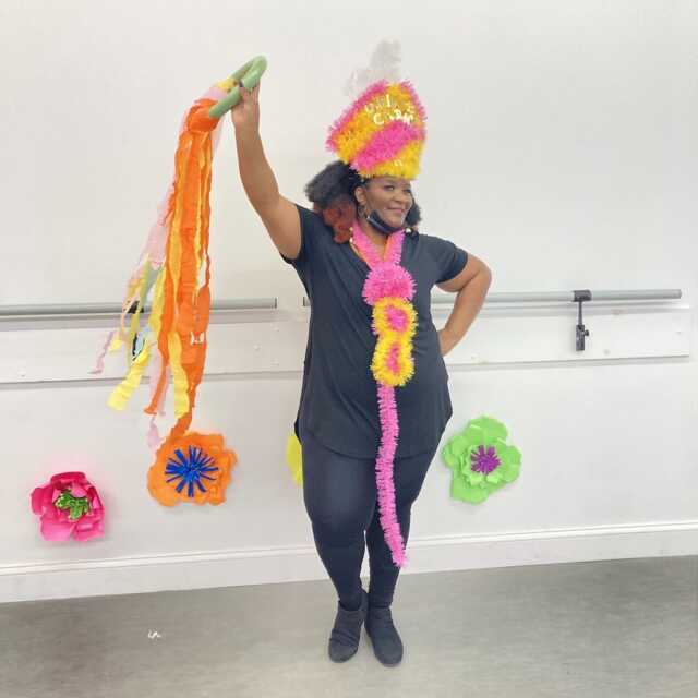 Woman with bright orange and yellow hat holding long ribbons in a dance studio.