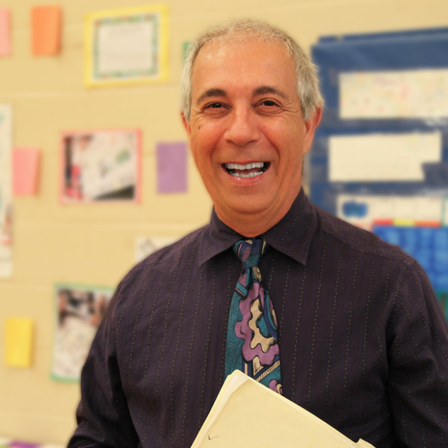 White man with short gray hair with a bright smile standing in front of a school bulletin board and holding a manilla folder.