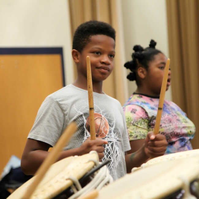 A middle school student in a gray t-shirt stands on stage smiling and holding drumsticks in a ready position behind large Japanese drums. Another student can be seen in the background.