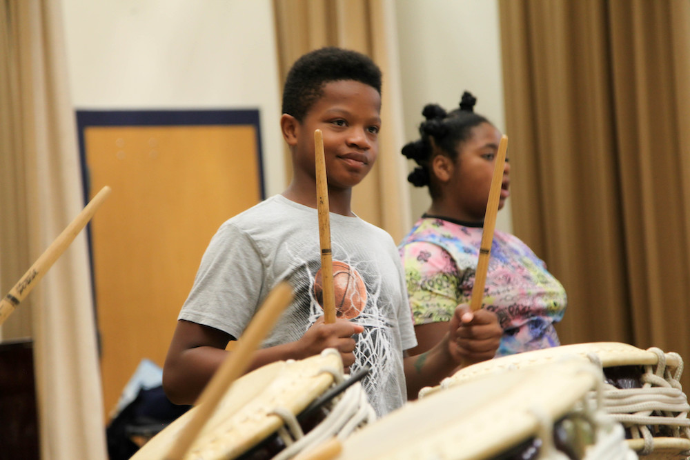 A middle school student in a gray t-shirt stands on stage smiling and holding drumsticks in a ready position behind large Japanese drums. Another student can be seen in the background.