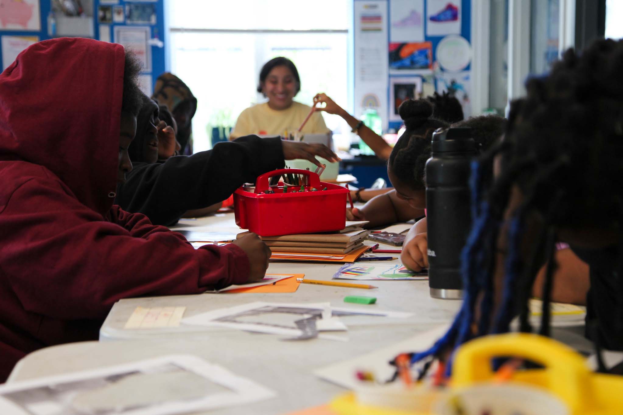 Ms. Nadia sits smiling at the end of a long table surrounded with students working on art projects. There is a red bin sitting in the middle of the table and students reaching inside of it for art supplies.