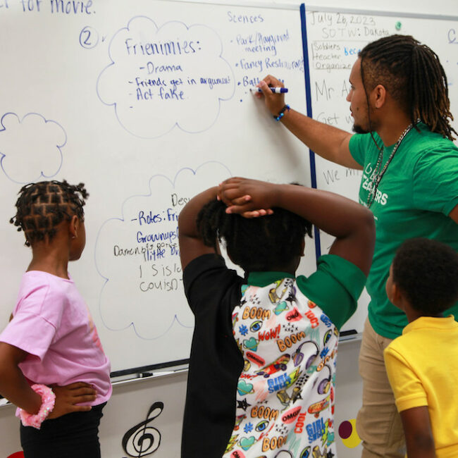 A teacher and students gather around a whiteboard and write out details of scenes for a student-created play.