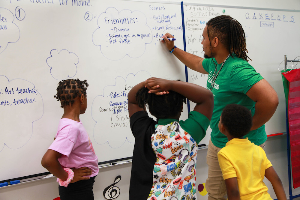 A teacher and students gather around a whiteboard and write out details of scenes for a student-created play.