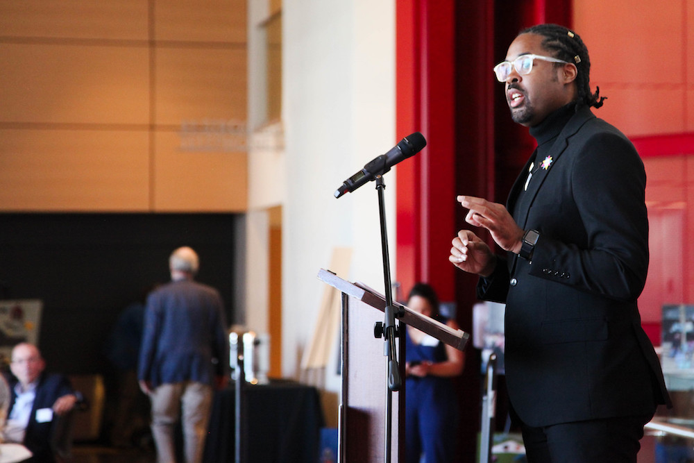 A man in a black suit with crystal-framed glasses speaks at a podium in front of red velvet curtains. People can be seen in the distance, standing at the right of the stage.