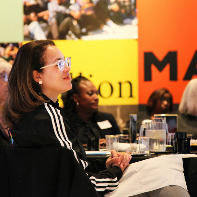 Side view of Sheelagh Allston seated at a table in a crowded room and looking ahead. She is wearing a black track jacket with white stripes and smiling.