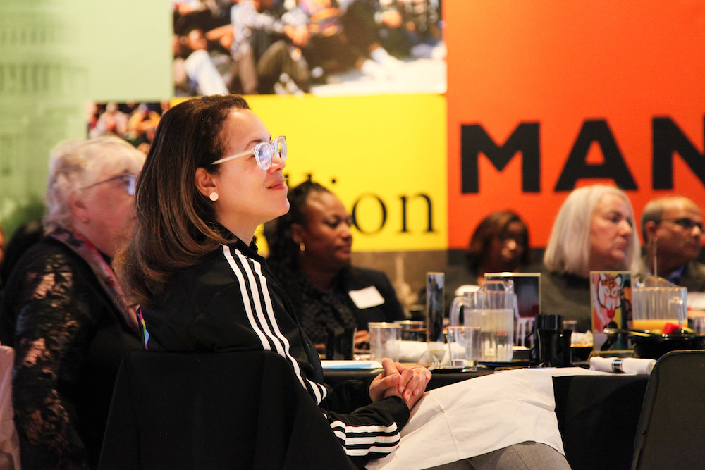 Side view of Sheelagh Allston seated at a table in a crowded room and looking ahead. She is wearing a black track jacket with white stripes and smiling.
