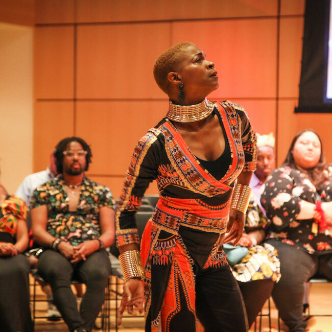 A woman stands mid-dance in front of a seated crowd. She's dressed in a gold and navy full-length leotard, draped in orange and gold printed fabric, and wearing a tall, gold collar necklace.