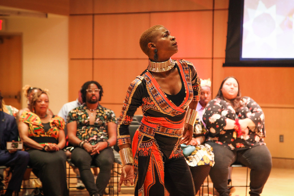 A woman stands mid-dance in front of a seated crowd. She's dressed in a gold and navy full-length leotard, draped in orange and gold printed fabric, and wearing a tall, gold collar necklace.