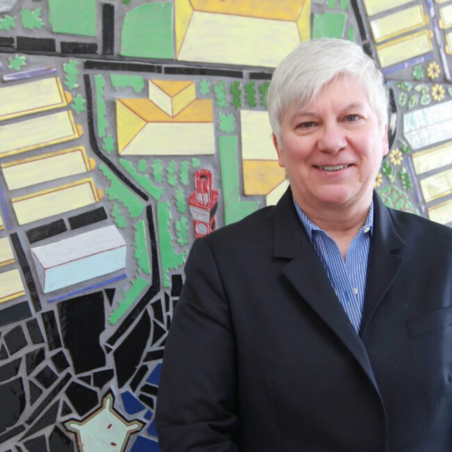 A woman wearing a blue collared shirt and dark blazer smiles. In the background is a colorful, geometric mosaic of spring green, black, and yellows.