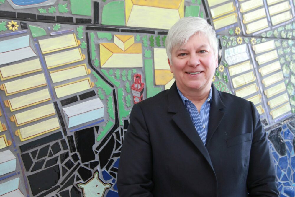 A woman wearing a blue collared shirt and dark blazer smiles. In the background is a colorful, geometric mosaic of spring green, black, and yellows.