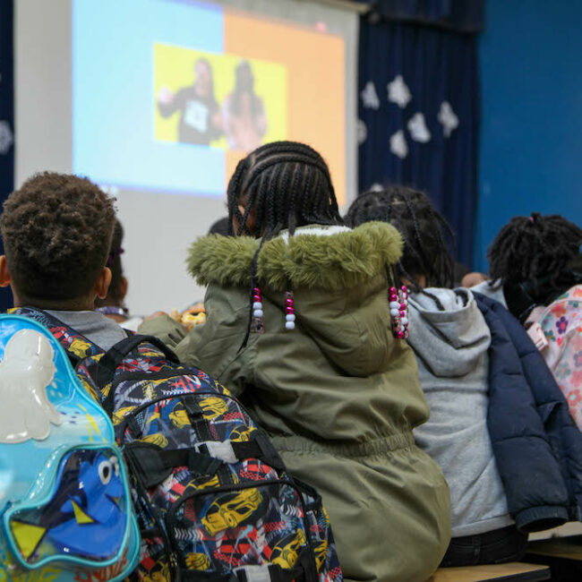 Students bundled in winter coats and wearing backpacks, sitting very close together at a cafeteria table. They are watching Blacktastic on a large screen at the front of the room.