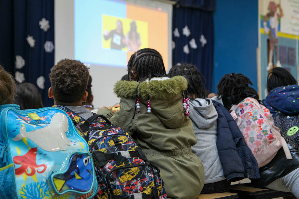 Students bundled in winter coats and wearing backpacks, sitting very close together at a cafeteria table. They are watching Blacktastic on a large screen at the front of the room.