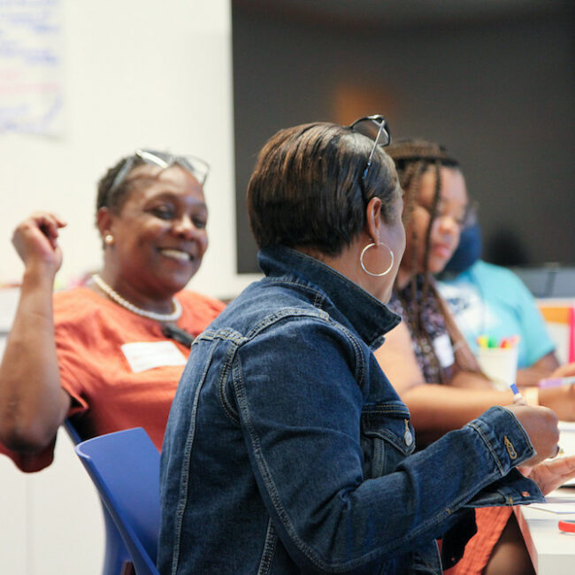 School principals sit around a long white table covered in scissors and different papers. One principal is turned and smiling. Her glasses are resting on the top of her head.