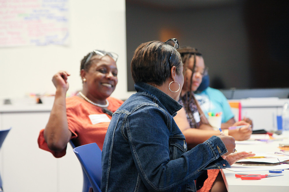 School principals sit around a long white table covered in scissors and different papers. One principal is turned and smiling. Her glasses are resting on the top of her head.