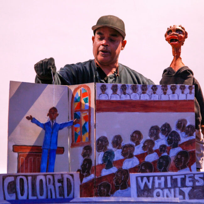 Schroeder Cherry, dressed in all black and wearing a blaseball hat, stands behind a large folded cardboard scene with one of his handmade puppets. In the scene, a preacher in a blue suit, arms outstretched above the word, "colored," faces rows of seated individuals in a full church above the words, "whites only."